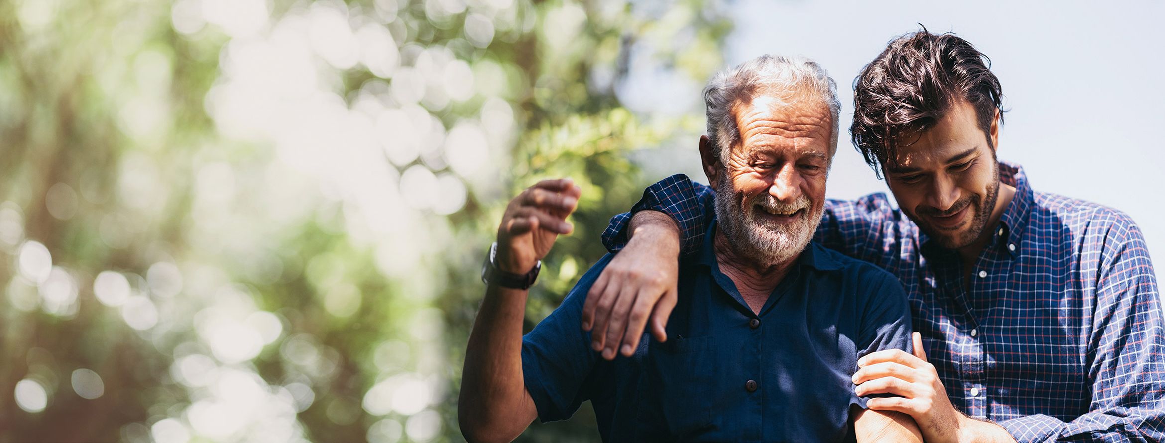 A father and son duo in each other's embrace outside laughing in the sunny weather.